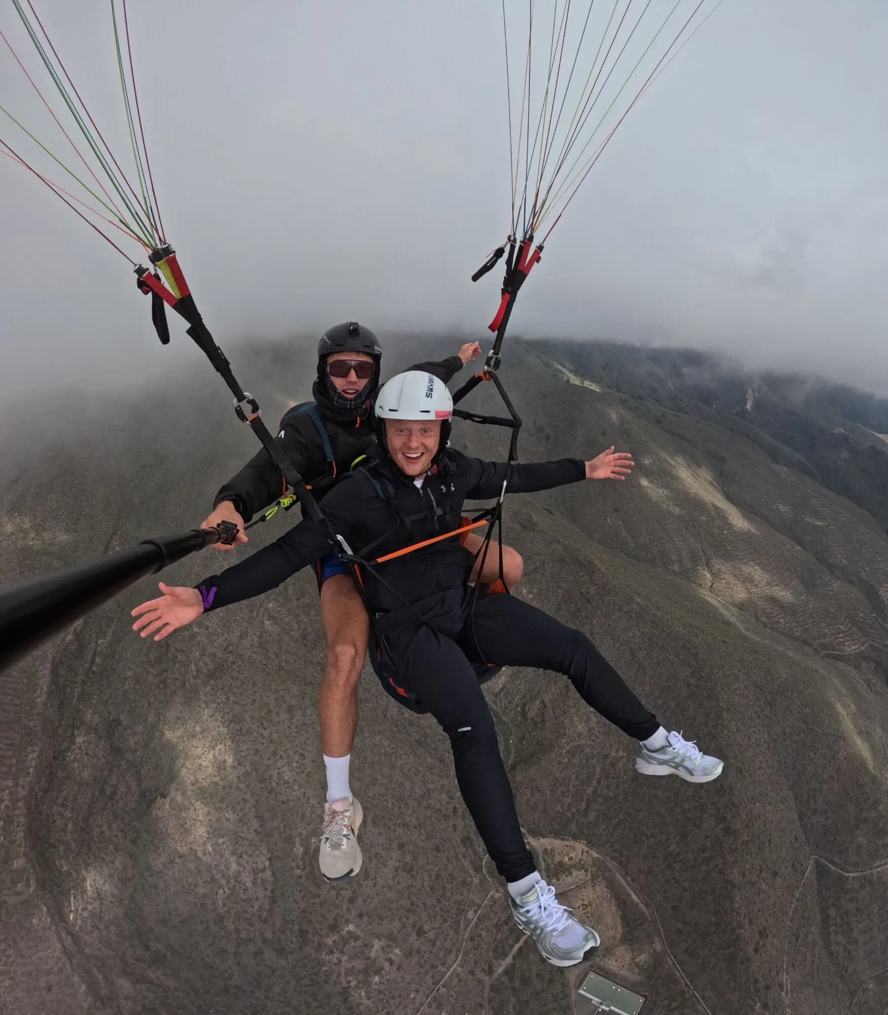 Pilot and passenger grinning under the wing above a cloudy mountain ridge